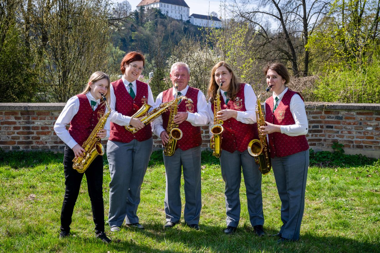 Fünf Personen in roten Westen und grauen Hosen stehen auf einer Wiese und halten Saxophone. Hinter ihnen befindet sich eine Ziegelmauer und eine Burg in der Ferne.