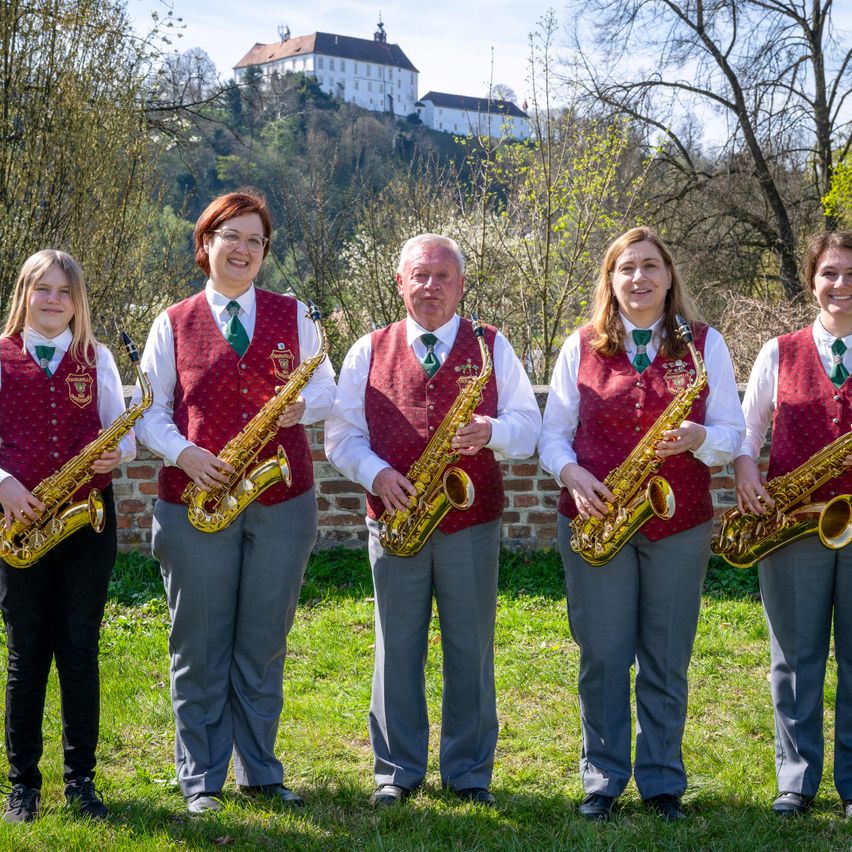 Fünf Personen in identischen roten Westen posieren mit Saxophonen vor einer Ziegelmauer. Dahinter ist ein Hügel mit einem Schloss und Bäumen zu sehen.