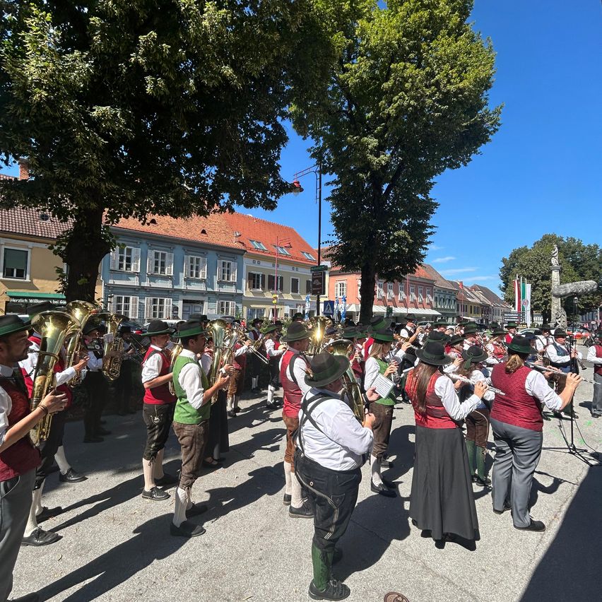 Eine Brassband spielt auf einem Stadtplatz, wobei die Musiker traditionelle Kleidung tragen. Die Band ist von Zuschauern umgeben, und Bäume und Gebäude sind im Hintergrund zu sehen.