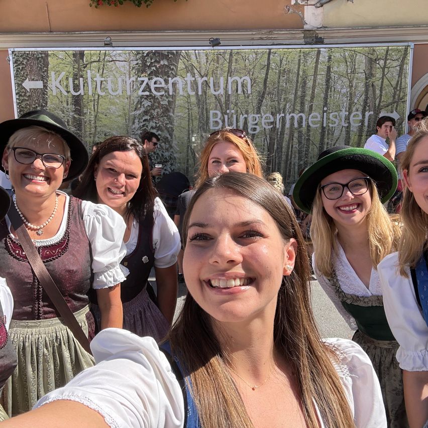Eine Gruppe von Frauen, die vor einem Kulturzentrum Burgermeister-Schild mit Bäumen im Hintergrund lächelnd für ein Foto posieren.
