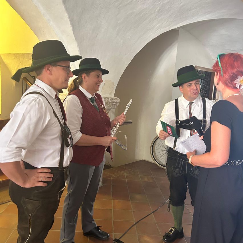 Vier Personen in traditioneller Kleidung, möglicherweise Musiker, unterhalten sich in einem schwach beleuchteten Raum mit gefliestem Boden. Eine Frau in Schwarz hält ein Papier, während ein Mann eine Flöte hält. Ein anderer Mann liest aus einem Büchlein.