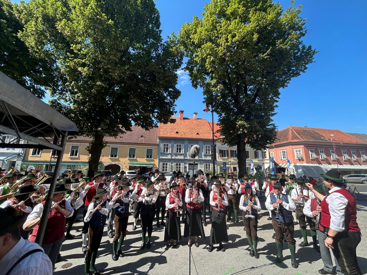 Eine Gruppe von Musikern in traditioneller Kleidung tritt auf einem Stadtplatz auf, umgeben von Bäumen und Gebäuden. Sie spielen verschiedene Instrumente unter einem blauen Himmel.