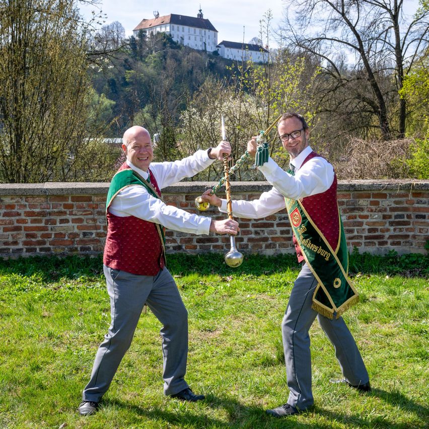 Zwei Männer in traditioneller deutscher Kleidung halten silberne Becher auf einer Wiese, mit einer Burg und Bäumen im Hintergrund.