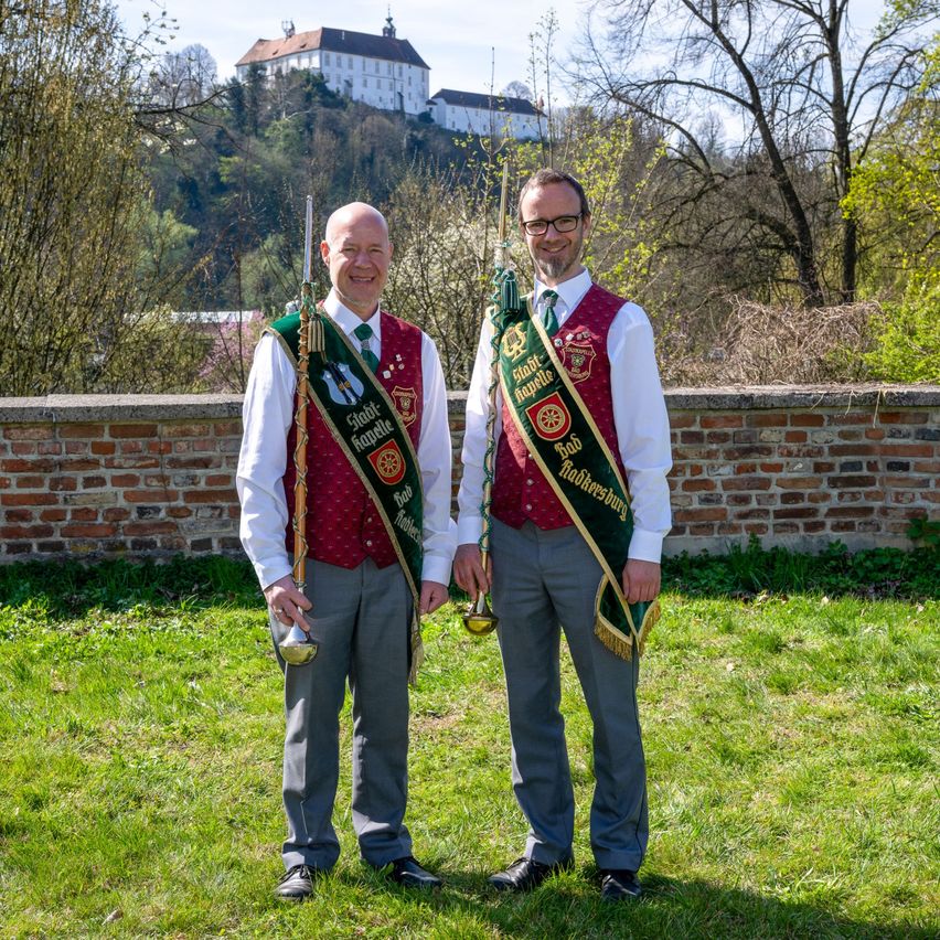 Zwei Männer in traditioneller Kleidung stehen auf einem Rasen mit einer Ziegelmauer im Hintergrund, lächeln und halten Trompeten. Ein Schloss ist auf dem Hügel hinter ihnen zu sehen.
