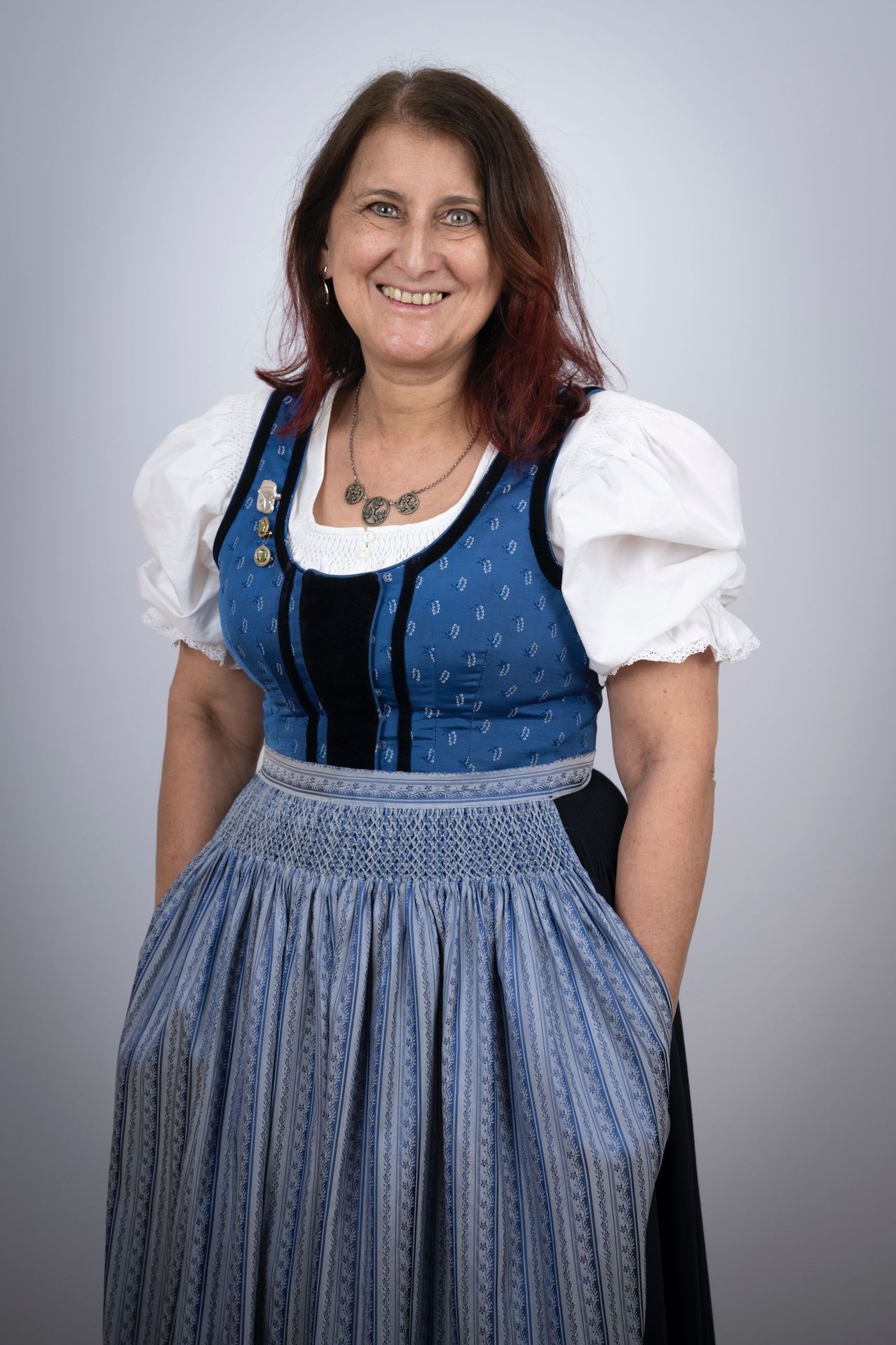 Eine Frau in einem blau-weißen traditionellen Kleid mit einer Halskette und Anstecknadeln auf dem Mieder, steht lächelnd mit den Händen in den Hüften.