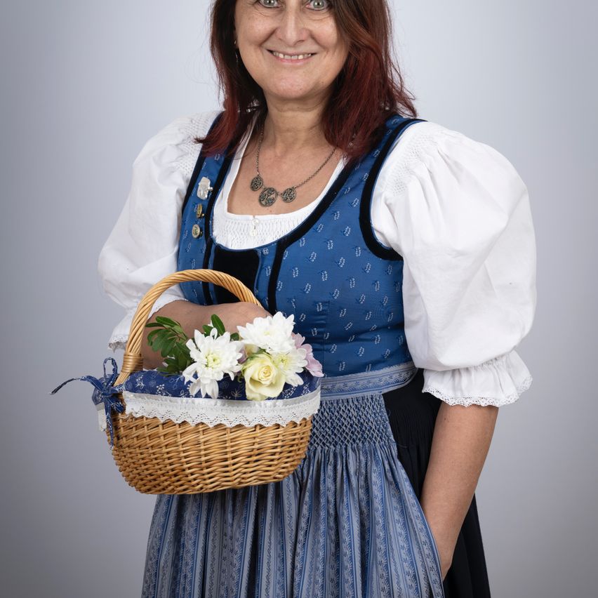 Eine Frau in traditioneller Kleidung, die einen Korb mit Blumen hält, lächelt für die Kamera.