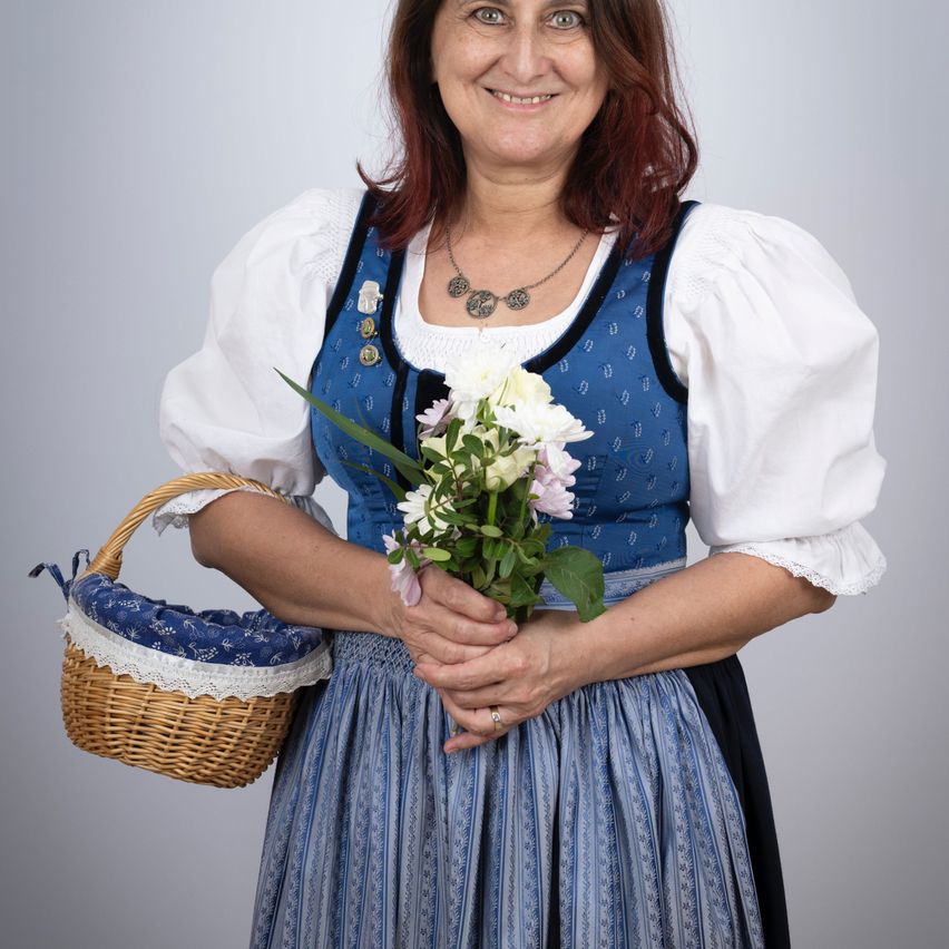 Eine Frau in einem traditionellen deutschen Kleid mit blauen und weißen Farben, das einen Blumenstrauß und einen Korb hält, lächelt für ein Foto.