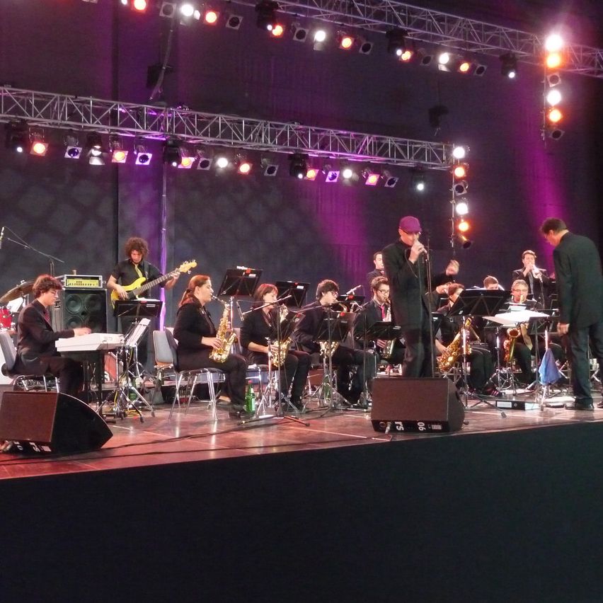 Eine Jazzband spielt auf einer Bühne mit lila Beleuchtung. Musiker spielen Instrumente wie Saxophone, Gitarre und Klavier. Ein Mann mit Mikrofon steht vorne.