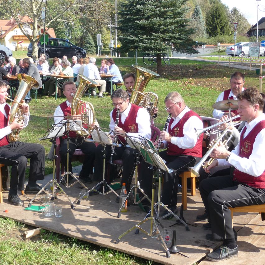 Eine Blechbläserband spielt im Freien, während im Hintergrund Menschen an Tischen sitzen. Bäume und geparkte Autos sind in der Ferne zu sehen.