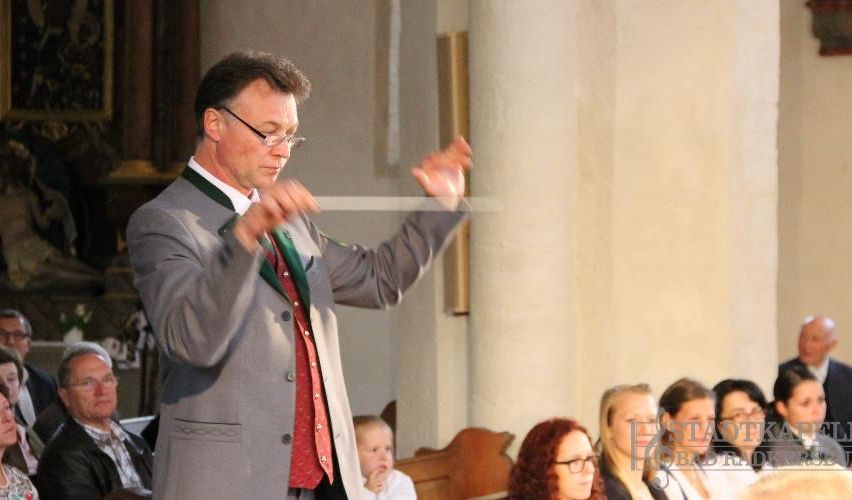 Ein Mann in einem grauen Anzug und einer roten Weste dirigiert eine musikalische Aufführung in einer Kirche. Im Hintergrund sitzen mehrere Menschen und lauschen aufmerksam.