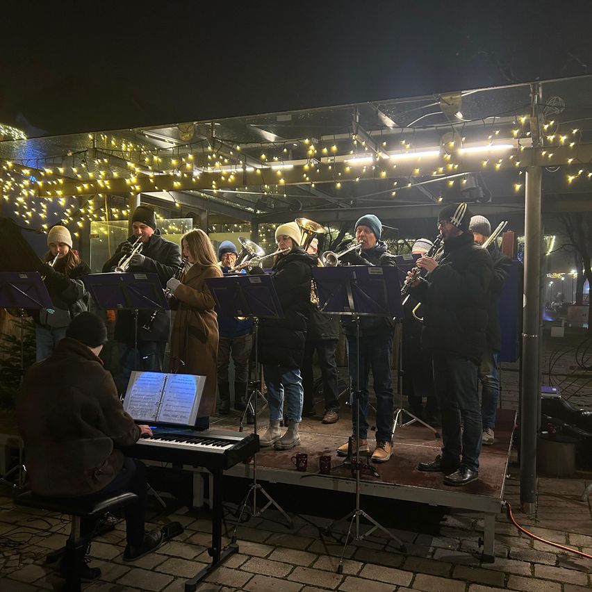 Eine Gruppe von Musikern spielt auf einer Bühne mit Saiteninstrumenten und einem Klavier, unter einem Baldachin mit Lichtern.
