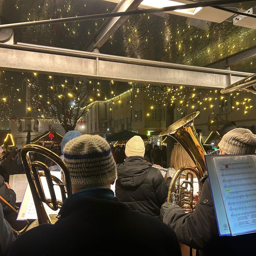 Eine Gruppe von Menschen spielt nachts Instrumente. Sie tragen Winterkleidung. Im Hintergrund sind Gebäude und beleuchtete Bäume zu sehen.