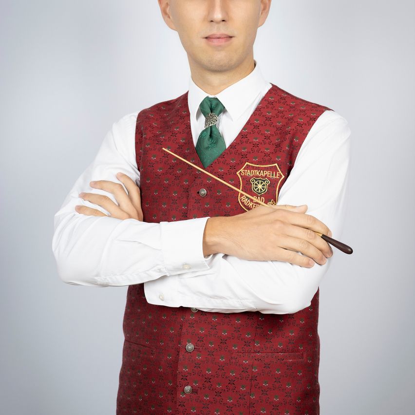 Ein Mann posiert mit verschränkten Armen in einem roten Weste mit weißen und grünen Mustern, einem weißen Langarmhemd und einer grünen Krawatte. Er hält einen Holzstab.