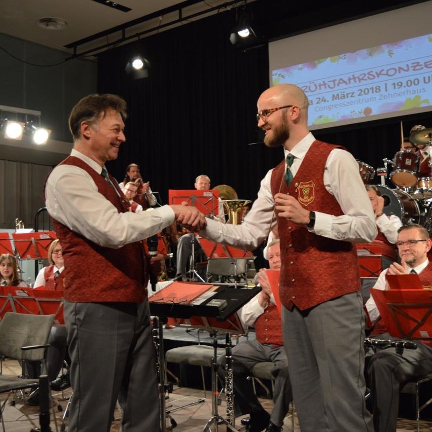 Zwei Männer schütteln sich die Hände auf einer Bühne mit einer Brass-Band hinter ihnen. Die Bühne hat Musikinstrumente und eine Projektionsleinwand mit dem Text '20-Jahrs-Konzert'.