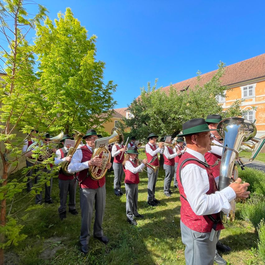 A brass band is performing in a park. They are playing brass instruments. The band members are wearing matching uniforms. The sun is shining brightly, and the sky is blue.