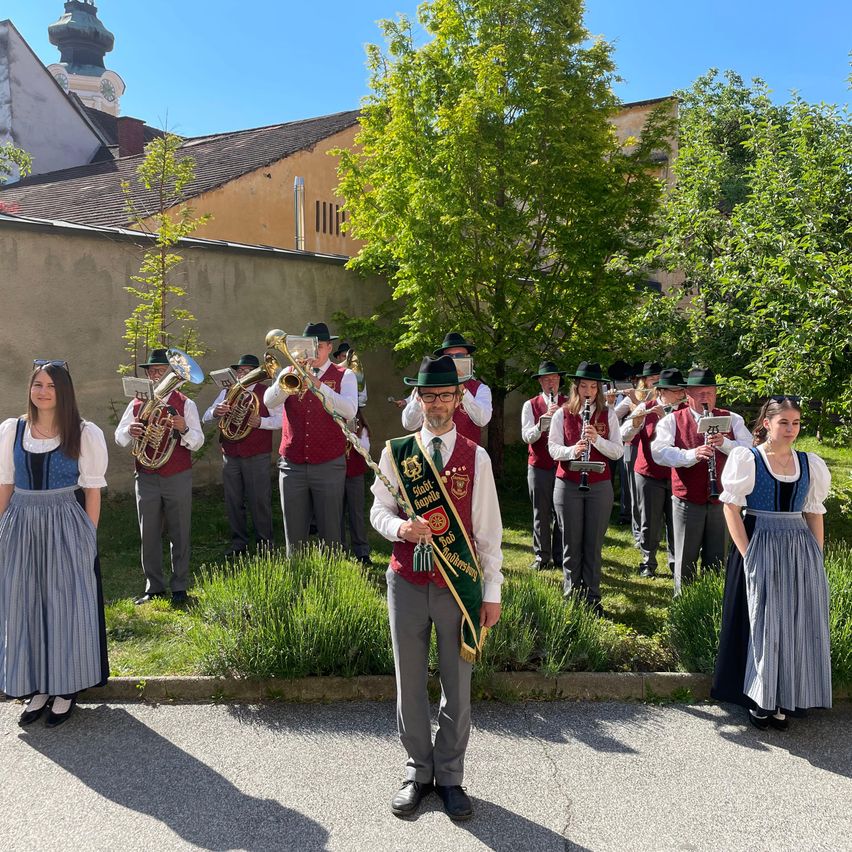 A band dressed in traditional attire stands in a line on the grass with one man holding a flag in front.