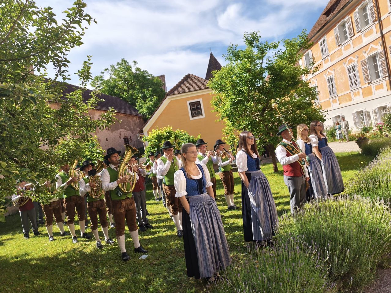 Eine Gruppe von Musikern in traditioneller Kleidung tritt in einem Garten auf, mit einem Gebäude und Bäumen im Hintergrund.