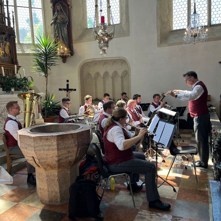 Eine Blechbläsergruppe übt in einer Kirche. Ein Dirigent steht und gestikuliert, während Musiker Trompeten, Posaunen und Tuben spielen. Die Kirche hat Buntglasfenster und eine Statue von Maria.