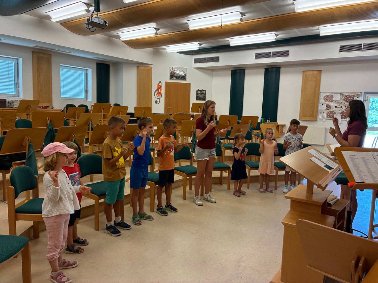 A group of children in a music room, standing in front of a woman who is likely their teacher, with musical instruments and a piano in the background.