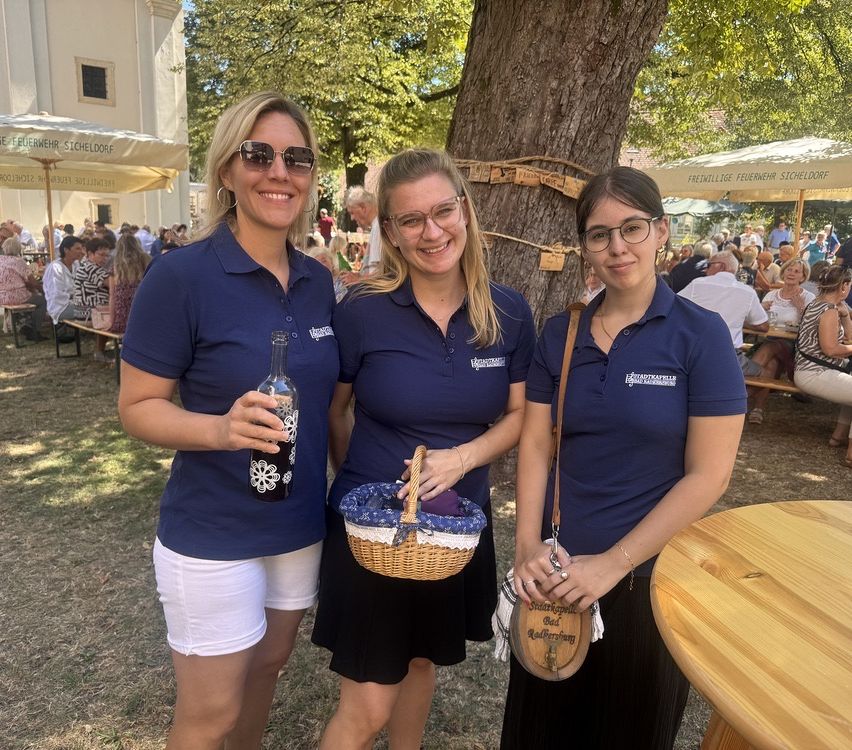 Drei Frauen in passenden blauen Poloshirts stehen in einer Außenumgebung, lächeln und posieren für ein Foto. Eine hält eine Flasche, eine andere einen Korb und die dritte eine Handtasche. Hinter ihnen ist ein Baum mit einem Seil und Notizen zu sehen, und im Hintergrund sitzen Menschen an Tischen unter Schirmen.