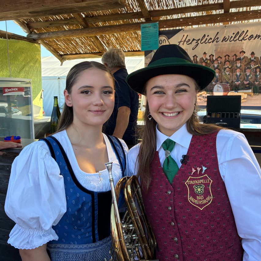 Two young women smile for the camera. One holds a brass instrument. They wear traditional Bavarian attire. Behind them, a tent with a banner and a blurred figure.