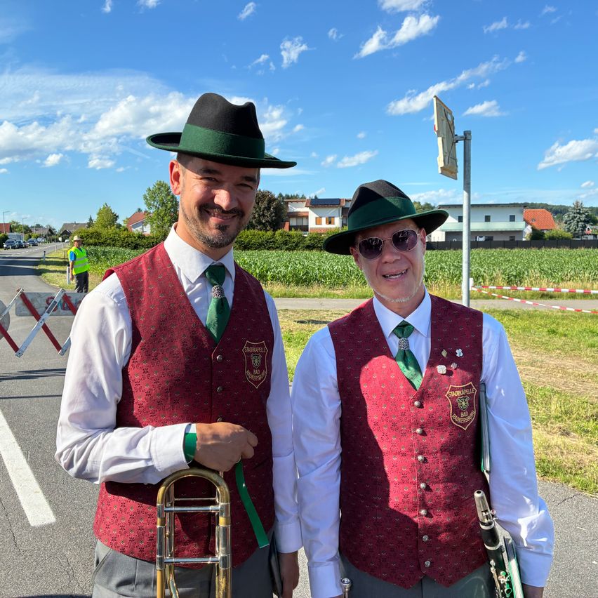Two men, dressed in maroon vests and green hats, stand on the side of a road with a trombone and a clarinet. A field and buildings are in the background.