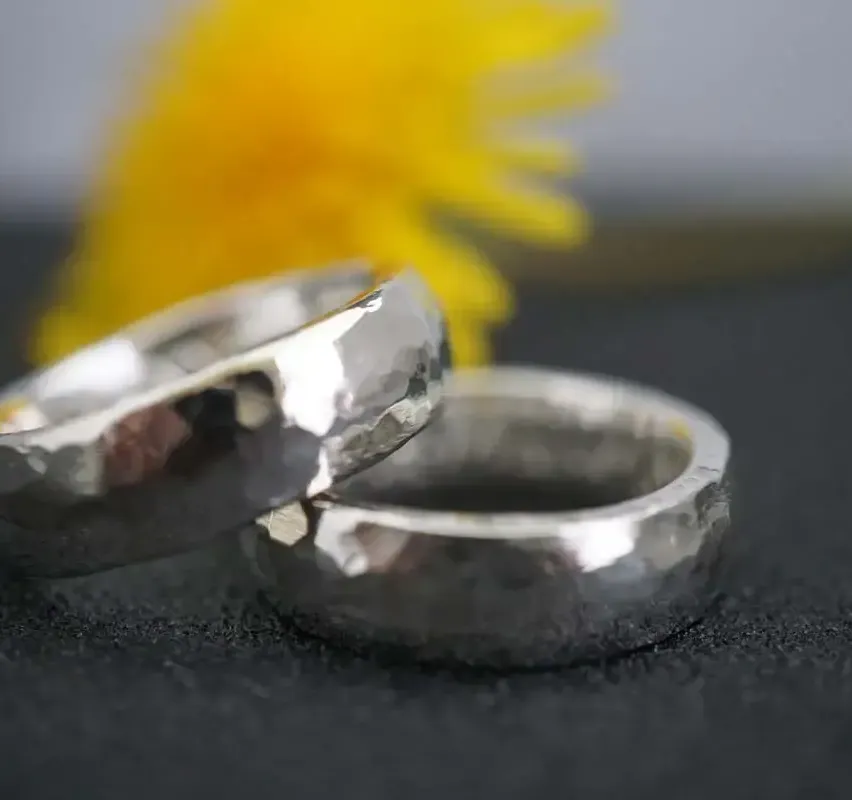 Zwei silberne Ringe mit strukturierter Oberfläche liegen auf einer schwarzen Unterlage mit einer gelben Blume im Hintergrund.