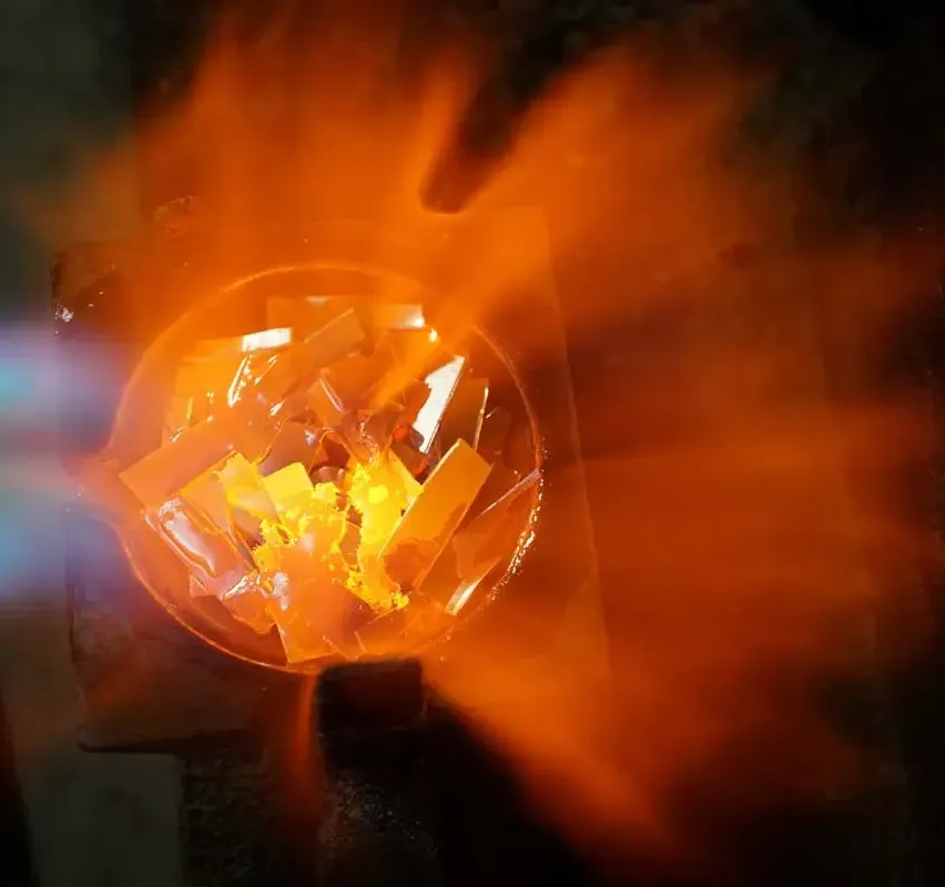 Eine Nahaufnahme eines Metallschüssels, gefüllt mit geschmolzenen Glasstücken, die unter einer hellen Flamme orange leuchten.