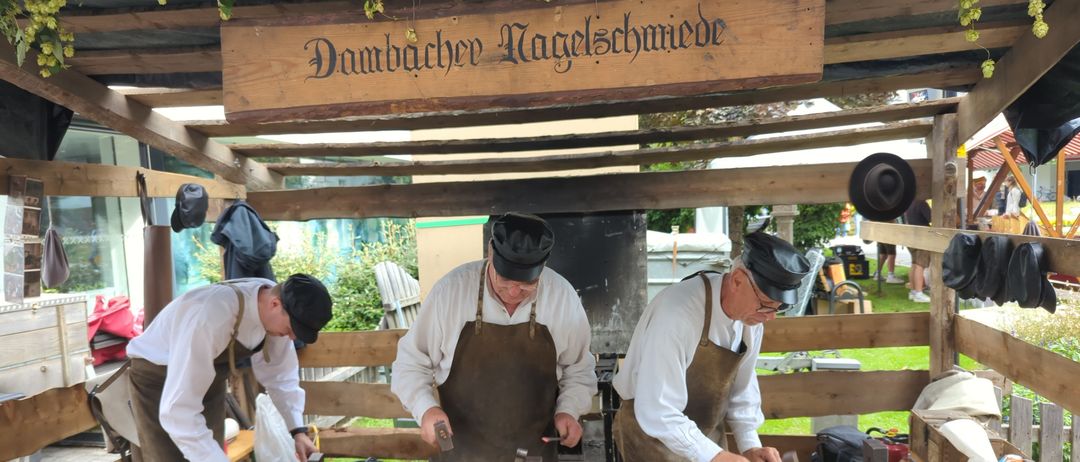 Drei Männer in Schürzen und Hüten arbeiten an Ambossen in einer Werkstatt mit einem hölzernen Schild über ihnen.