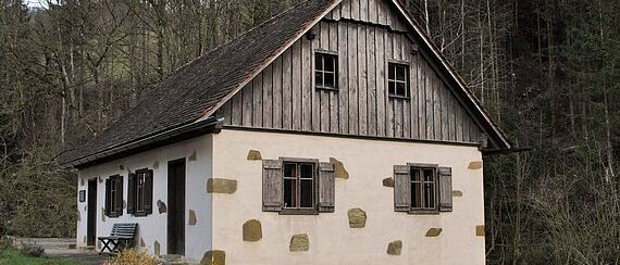 Ein kleines, rustikales Haus mit Holzschindeln und Steinakzenten steht in einer grasigen Lichtung, umgeben von einem dichten Wald.