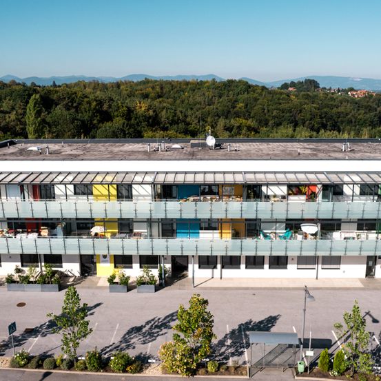 Luftaufnahme eines modernen Gebäudes mit mehreren Stockwerken, Glasbalkonen und Grünflächen davor. Das Gebäude ist von Bäumen und Bergen umgeben.