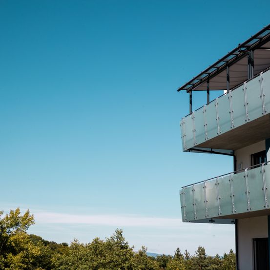 Ein zweistöckiges Gebäude mit einem Glasbalkon ist von Bäumen umgeben. Der Himmel ist klar und blau.
