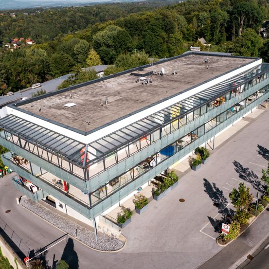 Luftaufnahme eines modernen Bürogebäudes mit flachem Dach und großen Glasfenstern, umgeben von Grünflächen und einem Parkplatz.