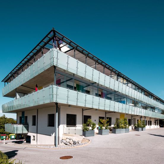 Modernes Apartmentgebäude mit Glasbalkonen und Dachterrasse. Große Fenster und Pflanzen zieren das Äußere. Bäume und ein klarer blauer Himmel im Hintergrund.