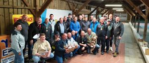 Eine Gruppe von Männern und Frauen posiert mit Medaillen und Trophäen lächelnd in einer Scheune. Ein Mann hält eine Box mit Gebäck, und ein weißer Kreis markiert den Boden.