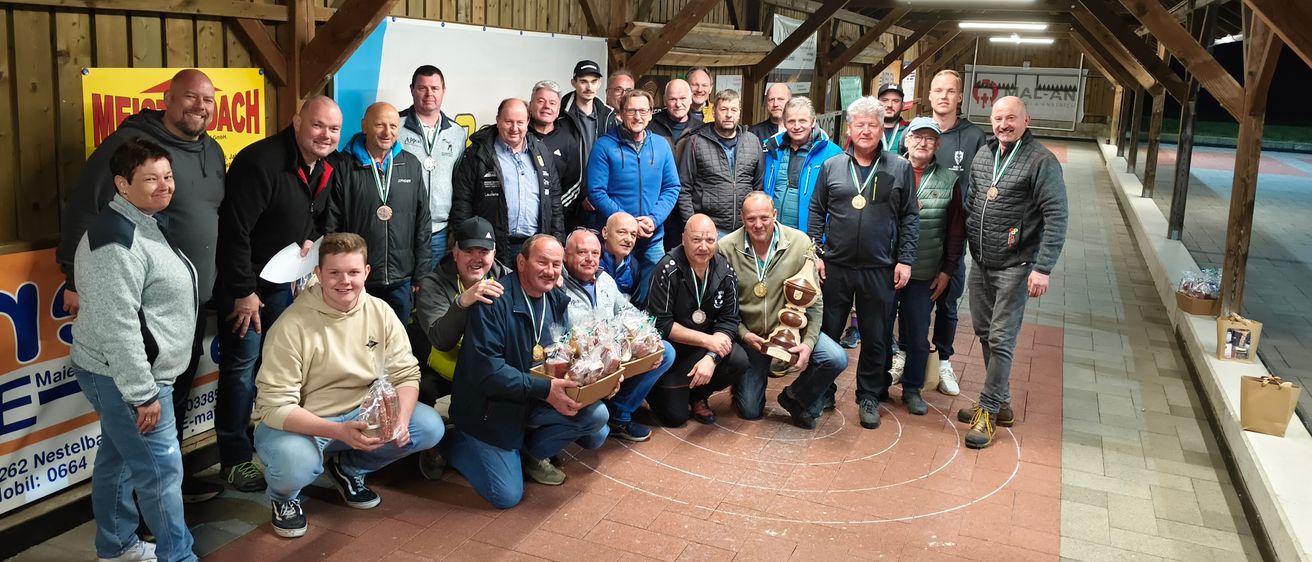 Eine Gruppe von Menschen posiert für ein Foto in einer Scheune, viele tragen Jacken und Medaillen, einige halten Geschenke. Der Raum hat Holzbalken und Lampen und ein Banner mit einem Logo im Hintergrund.