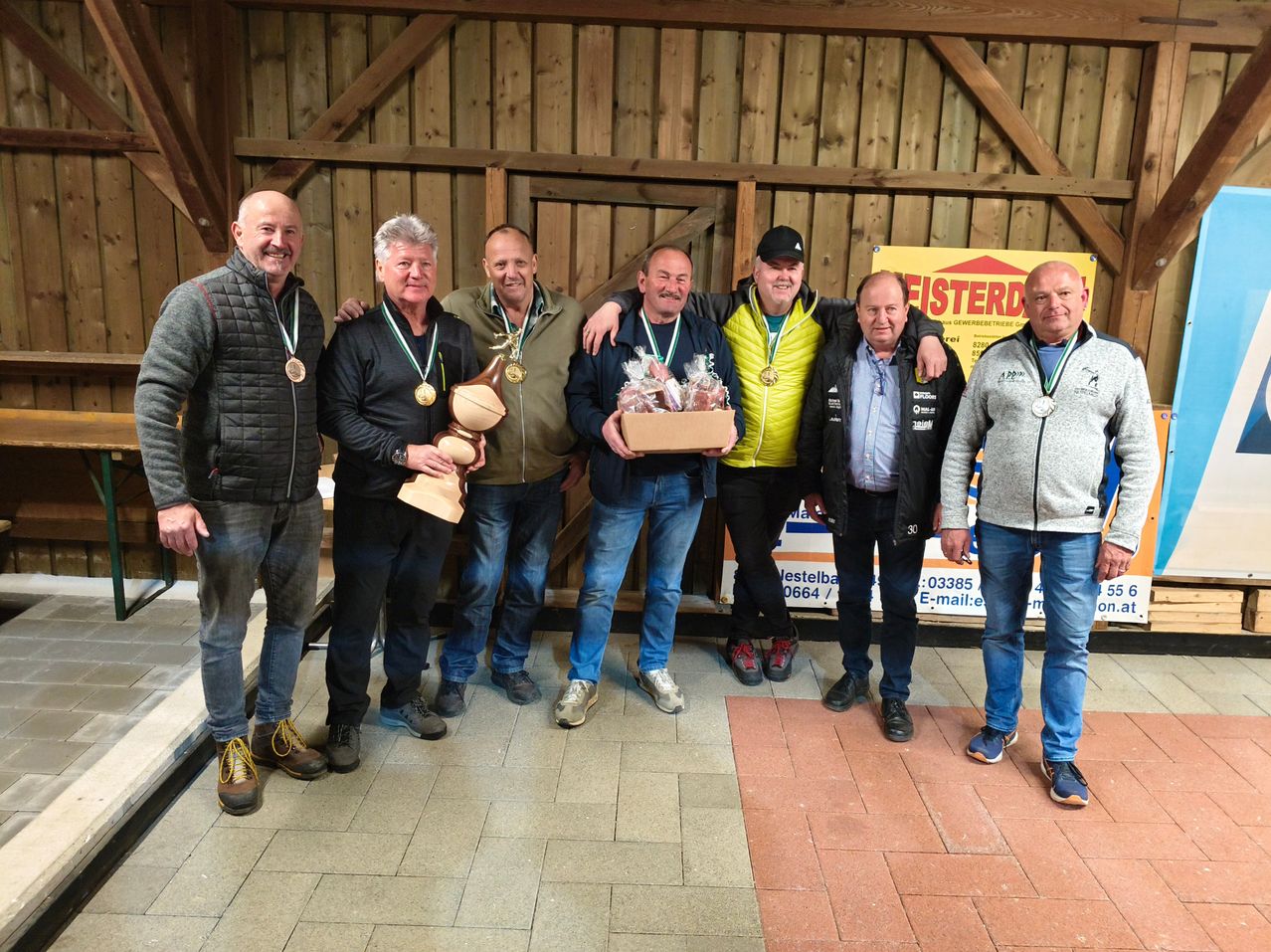 Sechs Männer stehen zusammen und halten Trophäen und Geschenke. Sie sind in einem Raum mit einer hölzernen Wand, und einer von ihnen hat ein Schild mit dem Wort 'FISTERD' darauf.