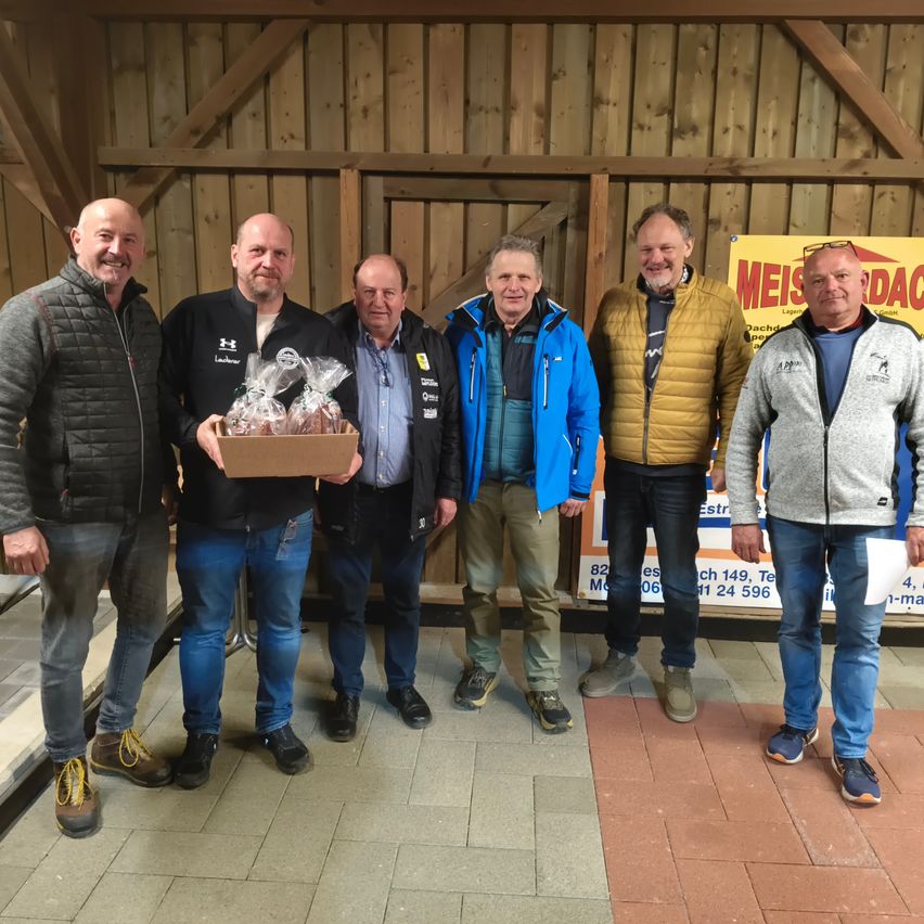 Fünf Männer posieren in einer Scheune, einer hält eine Schachtel mit eingewickeltem Brot. Hinter ihnen ist ein gelbes Schild. Sie tragen Jacken und Turnschuhe.