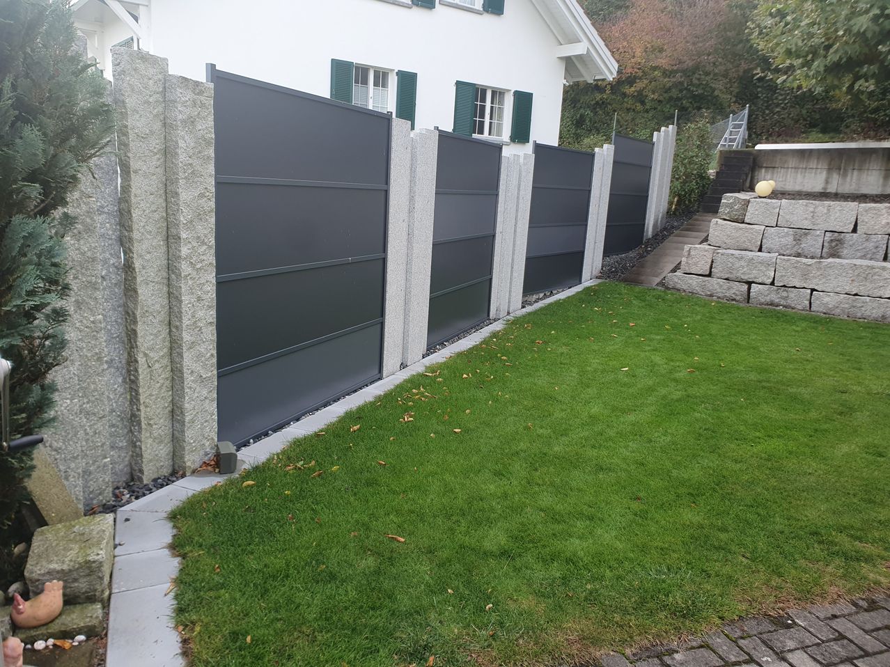 A white house with green shutters has a gray fence with multiple panels and a stone base. The fence is in front of a grassy yard with some fallen leaves. There is a small stone wall and a yellow ball on the side.
