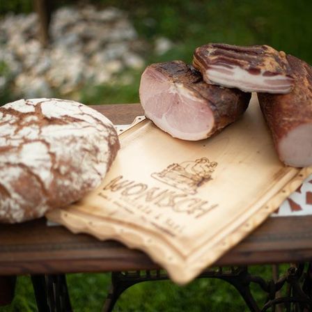Auf einem Holztablett auf einem Tisch befinden sich ein Laib Brot, zwei Scheiben geräucherter Schinken und eine Wurst. Der Hintergrund zeigt eine Rasenfläche.