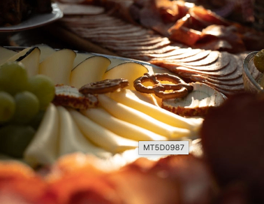 A close-up view of a charcuterie board with various sliced meats, cheeses, grapes, and pretzels arranged on a silver tray. The background is slightly blurred.