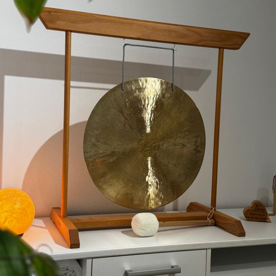 Ein goldener Gong hängt an einem Holzständer in einem Raum mit weißer Wand. Er wird von einem warmen Licht beleuchtet. Ein weißer Ball liegt auf dem Boden.
