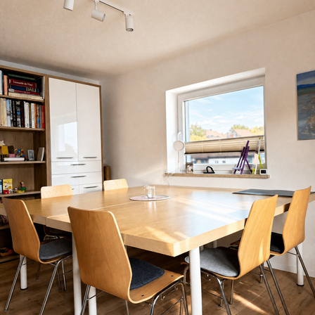 Ein hölzerner Esstisch mit vier Stühlen steht in einem Raum. Ein Schrank mit Büchern und Spielzeug steht links. Ein Fenster mit Jalousien und ein Gemälde befinden sich rechts.