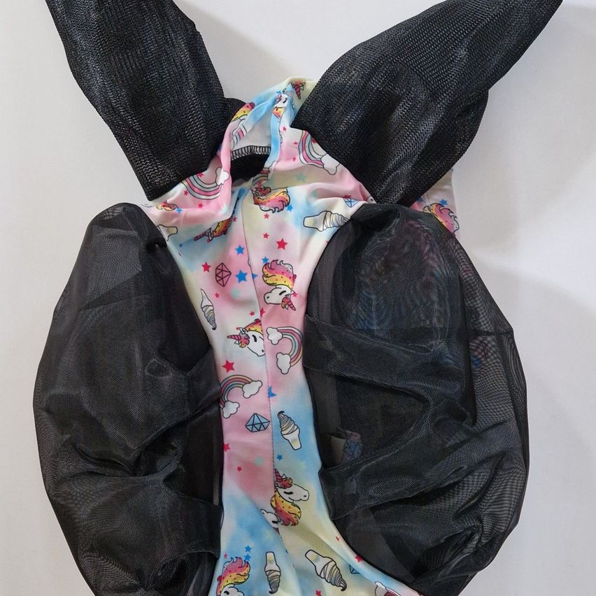 Ein rosa und blau bedruckter Stoff mit Einhörnern, Regenbogen und Eiscreme-Designs liegt auf einer weißen Oberfläche und ist von einem schwarzen Netzstoff mit zwei schwarzen ohrenähnlichen Abschnitten bedeckt.