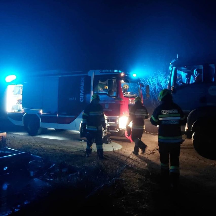 In der Nacht ist ein Feuerwehrauto mit eingeschalteten Lichtern am Straßenrand geparkt. Drei Feuerwehrleute in Uniform stehen darum herum.