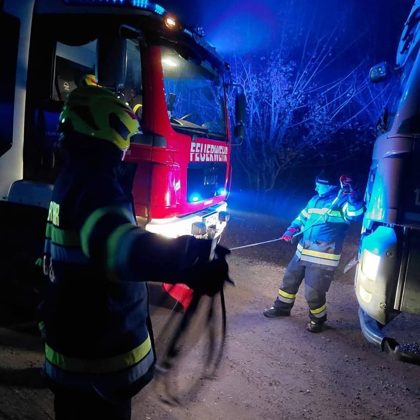 Zwei Feuerwehrleute stehen neben einem roten Feuerwehrwagen, einer hält eine Seil und der andere neben einem silbernen Fahrzeug. Die Szene ist nachts, mit den Lichtern des Trucks, die das Gebiet beleuchten.