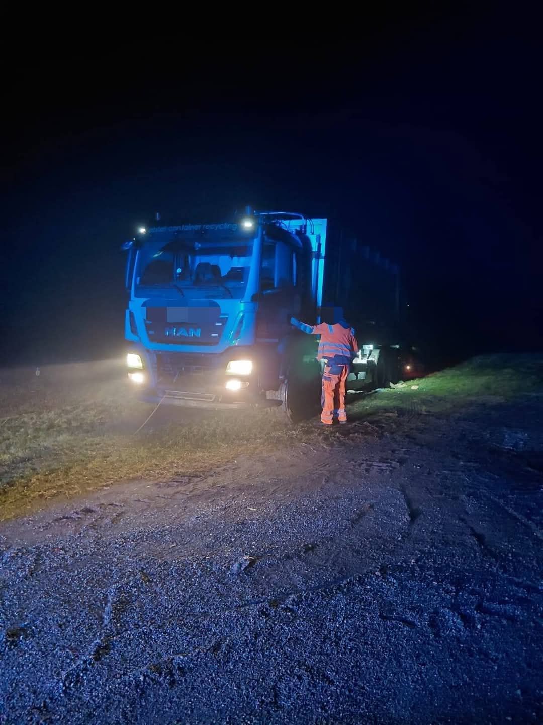 Ein MAN-Lkw steht nachts auf einer Kiesstraße, die Scheinwerfer sind eingeschaltet. Ein Arbeiter in einer orangefarbenen Sicherheitsjacke steht neben dem Lkw.