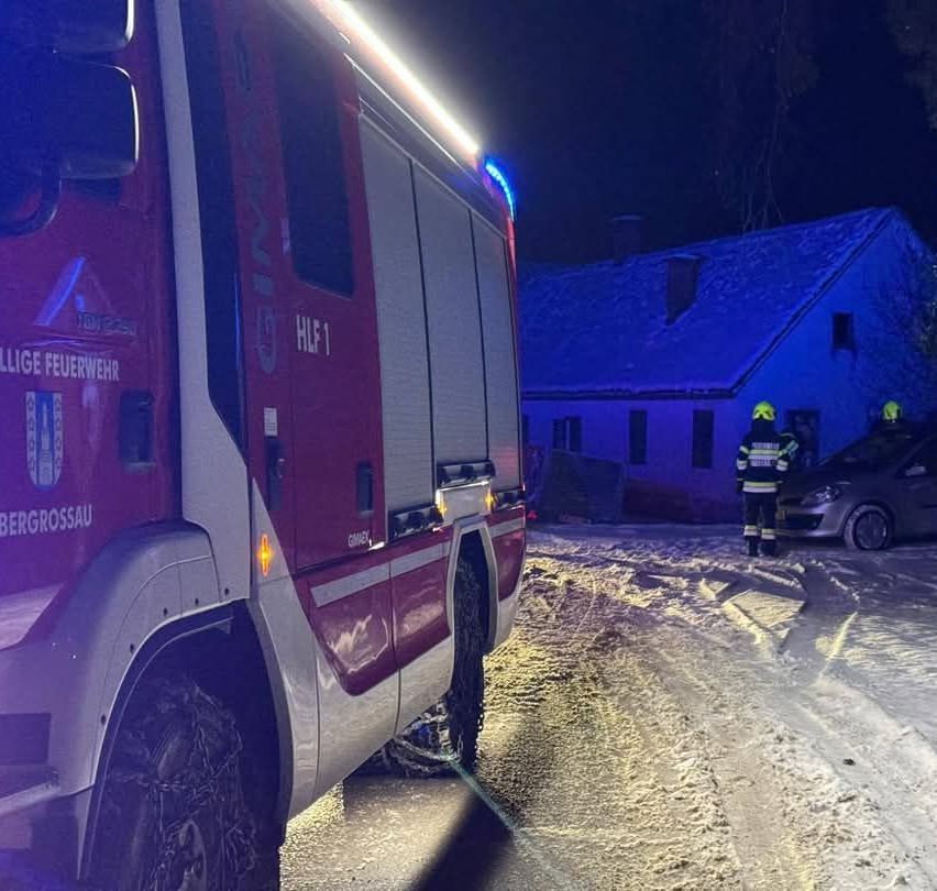 Ein Feuerwehrauto steht bei Nacht auf einer verschneiten Straße mit eingeschalteten Lichtern. Zwei Feuerwehrleute in gelben Helmen stehen bei einem Haus.