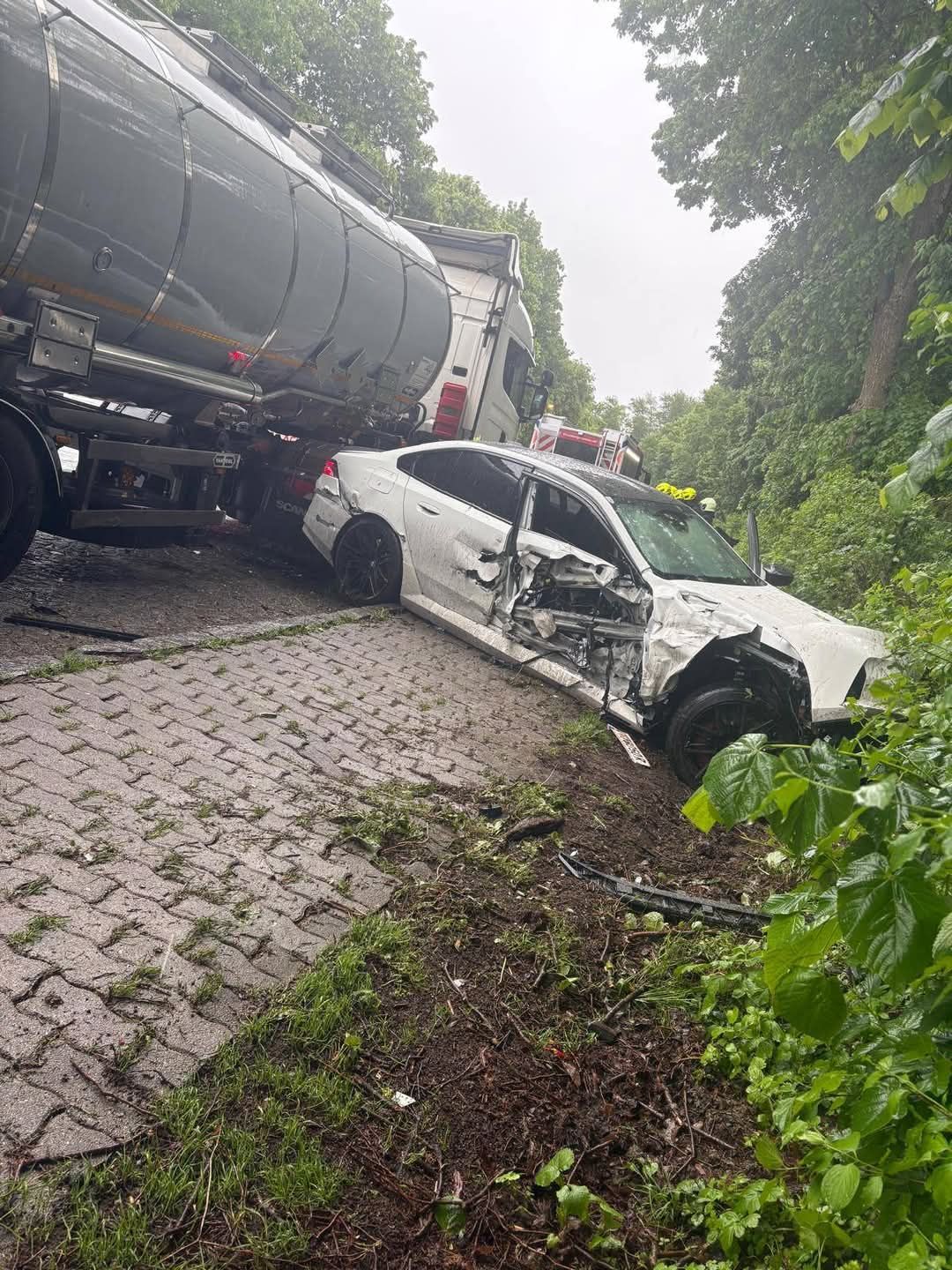A white car is wrecked and stuck next to a large truck. The car has sustained significant front-end damage. The scene is on a paved path, with a truck in the background.