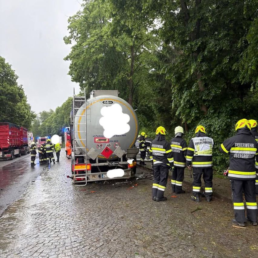 Firefighters in protective gear stand on a wet road, likely investigating a tanker accident. Other emergency vehicles and people are nearby.